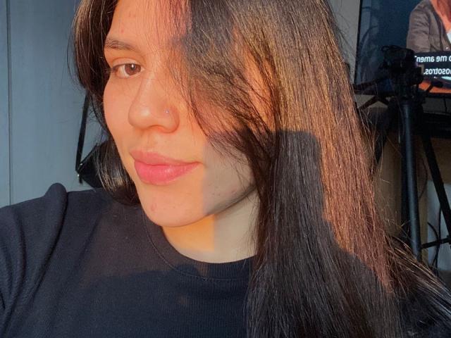 SophiaMarx with long brown hair, nose ring, and pink lips, wearing a black shirt, sunlit, shadow on right side, slightly smiling, soft light, relaxed expression, subtle freckles.