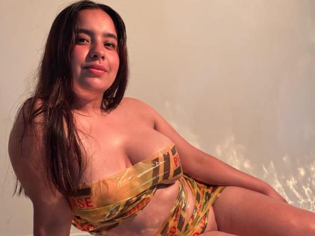 SammyTitss with long dark hair, tan skin, and a smile, wearing a yellow and orange striped bikini, showing her big tits and small ass. She's sitting with a relaxed look.