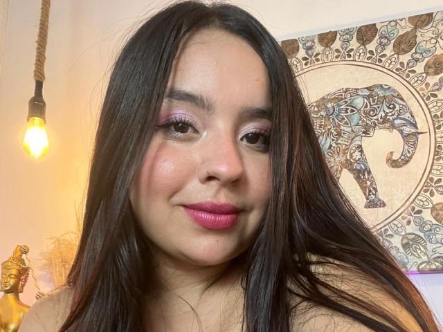 SammyTitss has long black hair, pink lipstick, and purple eyeshadow, with a subtle smile and a slightly flushed face. A lit bulb is in the background.