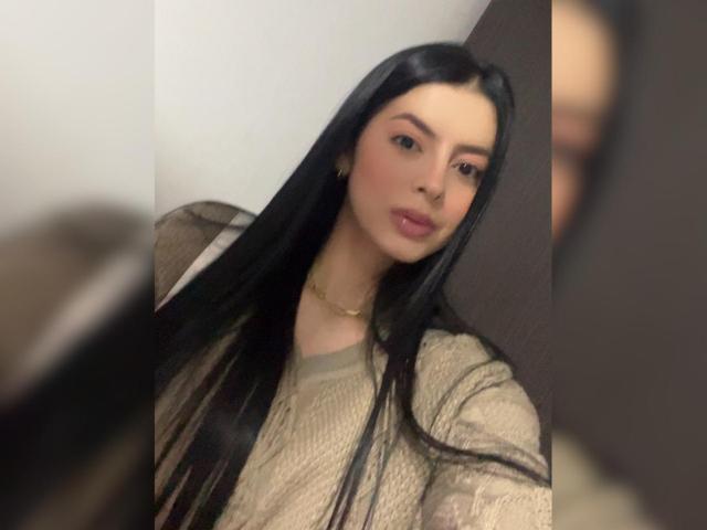 IsabellaJames with long black hair, wearing a sheer beige top, gold necklace, and pink lipstick, looking at a mirror with a soft, sexy expression.