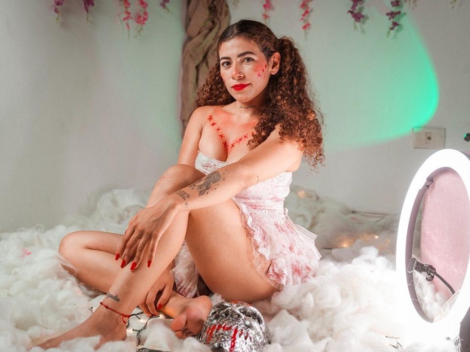 MiaBelly, con i capelli castano ricci, rossetto rosso, lingerie rosa in pizzo, tatuaggi, unghie rosse, seduta su un letto bianco e soffice, gambe incrociate, con una luce ad anello sulla destra e una luce verde sullo sfondo..
