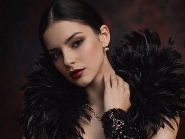 IsabellaJames with dark hair, red lipstick, and black feathered shoulders, wearing a sequined wrist cuff, looking down with a serious expression, touching her neck.