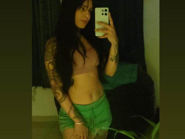Anflamel, avec de longs cheveux noirs, des tatouages sur les bras, un soutien-gorge rose, un short vert, tenant un téléphone, un selfie devant un miroir, montrant un ventre plat, éclairage vert dans la pièce, une plante verte en bas à droite..