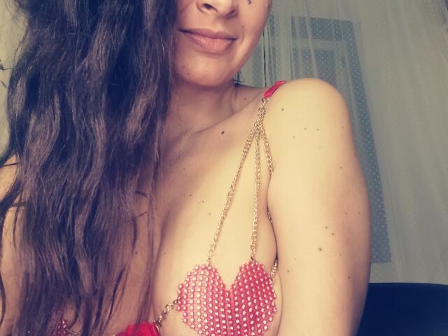 PassionX with wavy brown hair, smiling, showing pink beaded heart bra cups, covering her tits. Her skin is light with freckles. She's wearing a red strap.
