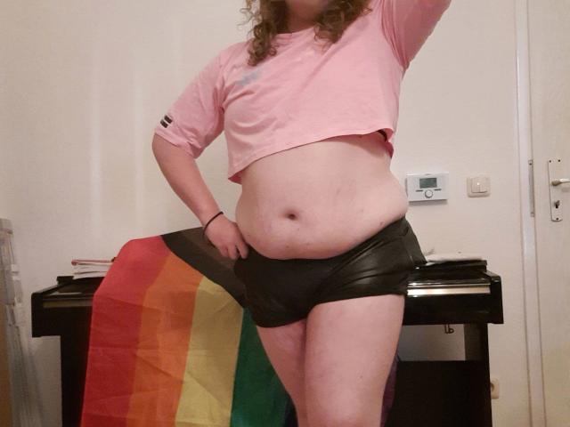 Juno-Mox, com cabelo castanho encaracolado, blusa cropped rosa e shorts de couro preto, segurando uma bandeira arco-íris, mostrando uma barriga um pouco saliente e pele clara..