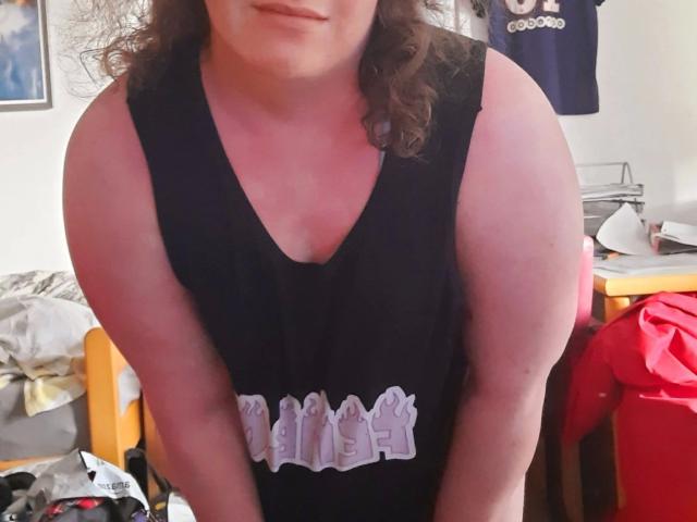 Juno-Mox-hot, eine mollige Person mit lockigem, braunem Haar, trägt ein schwarzes Tanktop, lehnt sich vor und zeigt ihre rosafarbenen Oberarme und Brust, lächelt leicht..