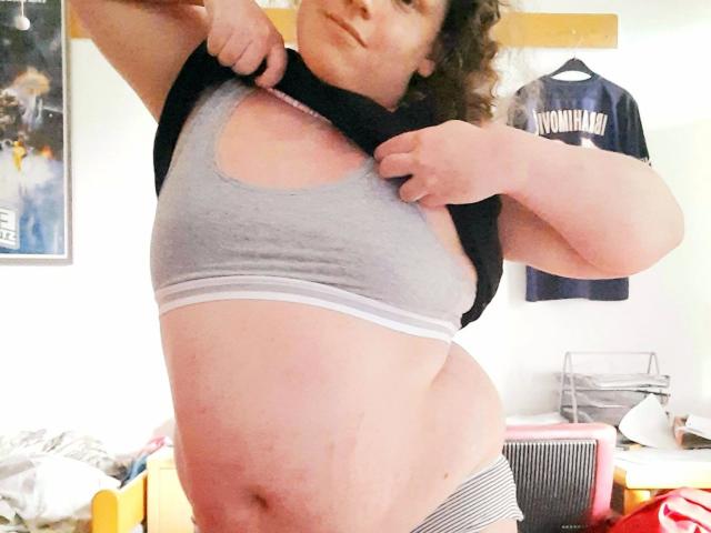 Juno-Mox, com cabelos castanhos cacheados, levantando uma camisa preta para mostrar seu sutiã esportivo cinza e uma barriga rechonchuda e pálida, com uma pequena hematoma. Ela está usando roupa íntima listrada..