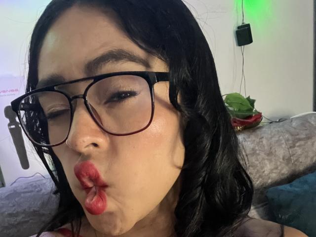StellaBeckker with black wavy hair, glasses, and red lips puckering, one eye closed, in a selfie. Her medium tits are partially visible. She has light skin and brown eyebrows.