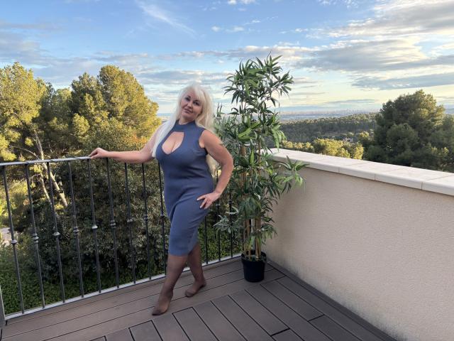 KamaliaSim with long blonde hair, blue cut-out dress, showing her tits, standing on a wooden balcony, hand on railing, potted plant by her side, smiling, wearing brown heels.