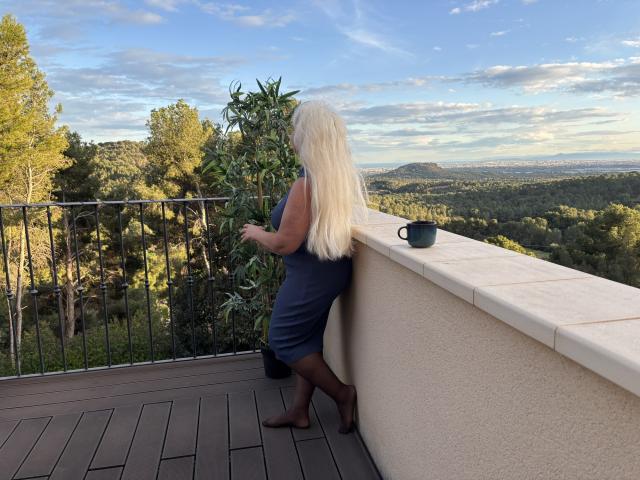 KamaliaSim with long, straight, platinum blonde hair stands on a wooden deck, facing a railing. She wears a navy blue, sleeveless dress and black tights. Her back is to the viewer. A black mug sits on the railing to her right. The background shows a lush, green forest with a clear, blue sky and scattered clouds. KamaliaSim's right hand rests on the railing while she looks out. The deck has a wooden texture, and the railing is black with vertical bars. The landscape is expansive and sunlit.