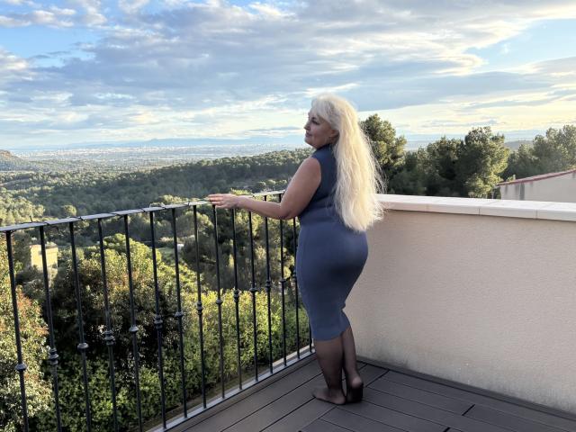 KamaliaSim stands on a wooden deck, facing a black iron railing. She has long, platinum blonde hair and wears a tight, sleeveless, grey dress that accentuates her curvy, full-figured physique. She is looking over her shoulder, with a slight smile. She is wearing black high heels. The background shows a lush, green landscape with trees and a distant horizon. The sky is partly cloudy with soft, blue hues. The image has a natural, outdoor setting with a clear, elevated view.
