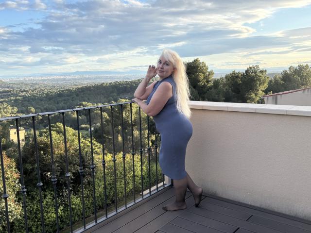 KamaliaSim with long blonde hair, blue dress hugging her big ass and small tits, standing on a balcony, hands near face, looking at camera, wooded valley in background.