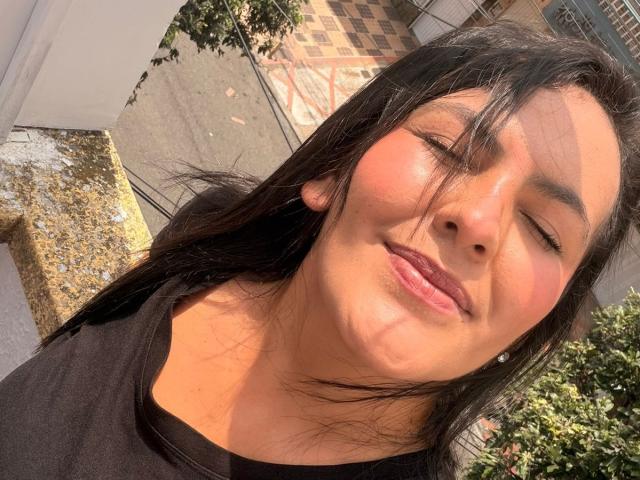 ScarlettOxford has medium brown skin, black straight hair, and is smiling with eyes closed, wearing a black top. Sunlight casts shadows on her face. Her lips are slightly parted.