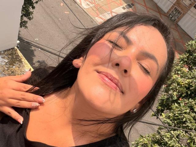 ScarlettOxford has sunlit, closed-eye face with black hair, pink blush, glossy lips, and a hand with glitter nails touching her neck. She's wearing a black top, with trees in background.