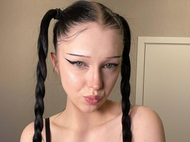 AmaiRin with puffy black twin braids, sharp winged eyeliner, glossy pink lips, and a wet, shiny forehead, wearing a black top, looking down with a pouty expression.