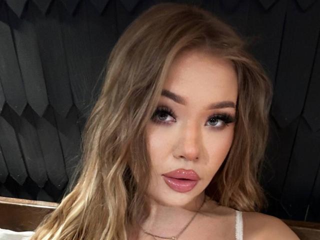 Close-up of StacyYellig. She has long, wavy blonde hair, full eyebrows, and heavy, dark eyeliner with long lashes. Her lips are glossy and slightly parted. She's wearing a white strap, and a thin necklace. The background is dark with a textured pattern. Her skin is fair and she has a slight, confident expression. The image is focused on her face, with soft lighting highlighting her features. She has a strong, attractive look.