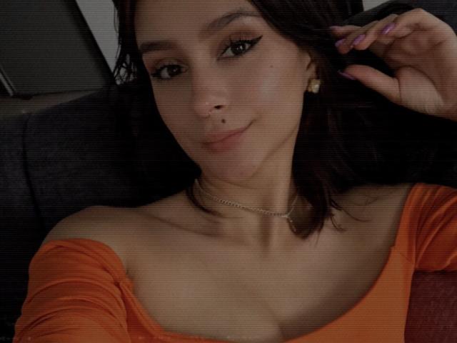 CamilaJolie with dark brown hair, light skin, wearing an orange off-shoulder top, smiling, purple nails, gold earrings, and a thin necklace, lying back, right hand touching hair.