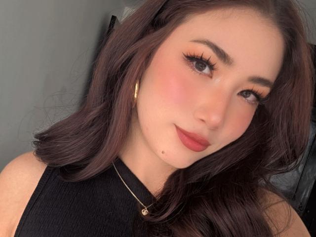 Stardustt has dark brown hair, full eyebrows, long eyelashes, pink blush, red lipstick, small beauty mark on her right cheek, black sleeveless top, and gold hoop earrings.