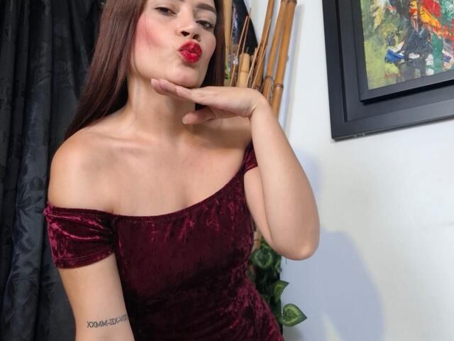 ShantalaKrisha with pouty red lips, dark brown hair, off-shoulder red velvet dress, hand on cheek, tattoo on left arm, standing in front of wall with framed painting.