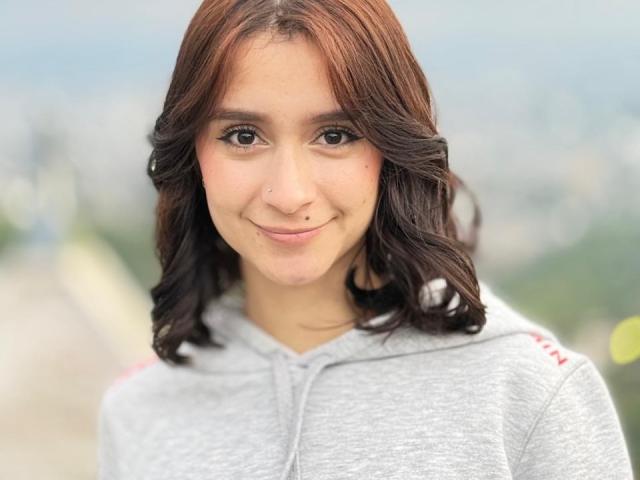 Close-up of CamilaJolie with shoulder-length, wavy brown hair. She has a subtle smile, brown eyes, and a small beauty mark on her right cheek. She wears a light gray hoodie with red stripes on the sleeves. Her skin is smooth and fair. The background is blurred, focusing attention on her face. The image uses natural light, highlighting her facial features. The overall composition is centered, with a slight upward tilt.