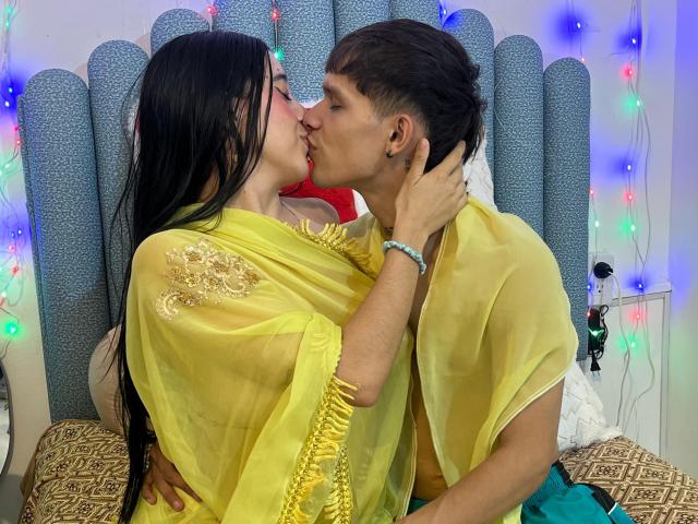 AnjaliRohan with long black hair, yellow sheer top, kissing a brown-haired guy, both topless, her hand on his neck, colorful lights in the background, bed with patterned sheets.