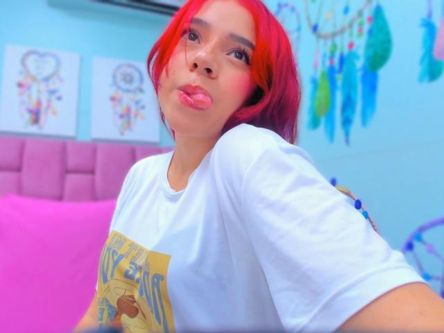 MiaLeroy, com cabelo ruivo, lábios curvados para baixo, camisa branca e travesseiro rosa, com a língua ligeiramente para fora, olhando para a câmera, em um quarto colorido com redes de sonhos nas paredes..