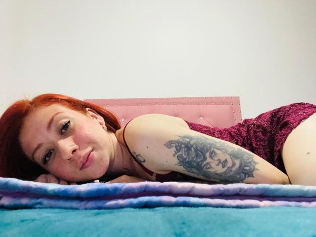 Valeriaaf with red hair, lying on a bed, pouty face, tattoo on right arm, wearing purple lace dress, looking at camera, bed has blue and purple blanket.
