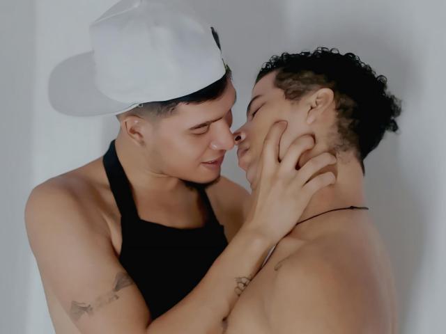 MaikolAndAndry, a muscular, shirtless man with short-cropped hair, wears a white baseball cap and a black halter. He gently holds the face of another MaikolAndAndry, who is also shirtless with curly hair and a thin black necklace. MaikolAndAndry's tattoos are visible on his right arm. They are close, eyes nearly touching, in an intimate moment. Soft, natural light highlights their skin tones. The composition emphasizes their emotional connection. The image is a close-up, focusing on their faces and upper torsos.
