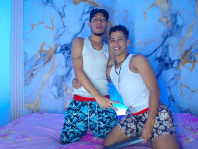 MaikolAndAndry, twee jongens, gekleed in witte tanktops en kleurrijke shorts, staan op een roze bed, waarvan er één een blauw-witte lichtstok vasthoudt en beiden glimlachen. MaikolAndAndry staat links..