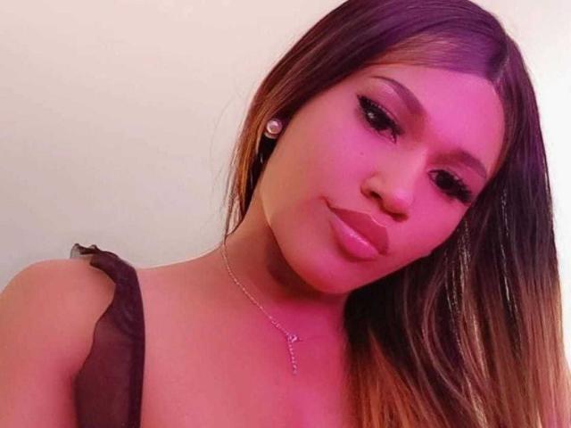 KloeDollyHot with pinkish-purple lighting, brown hair, and shiny lips, wearing a black strap top, and a necklace with a small cross, giving a pouty expression.