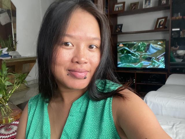 NellahHottie with black hair, green polka-dot sleeveless top, and a lip piercing, smiling at the camera in a living room. TV in background shows a green nature show.