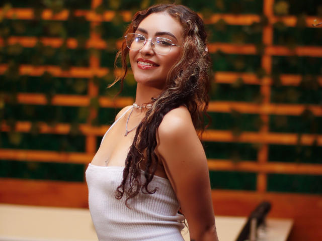 AdaraAngel with curly brown hair, glasses, white tank top, smiling, showing tits, necklace, and red lipstick, standing, looking over shoulder, in a warm-lit, wooden, green-walled room.