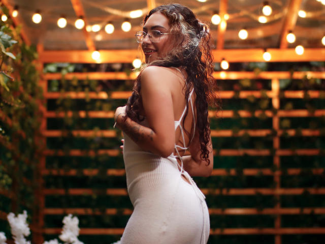 AdaraAngel with curly brown hair, glasses, and a tight white dress, showing her ass, smiling, hands on her back, string lights in the background.