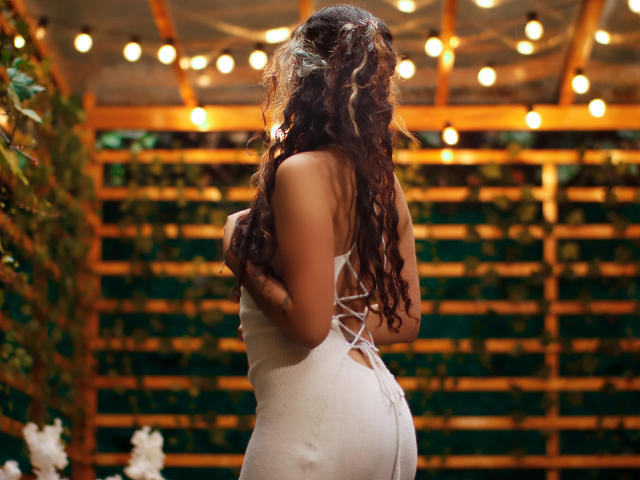 AdaraAngel with curly brown hair, wearing a white, lace-up, bodycon dress, showcasing her ass and back, standing under warm, string lights, in a wooden, green, and leafy setting.