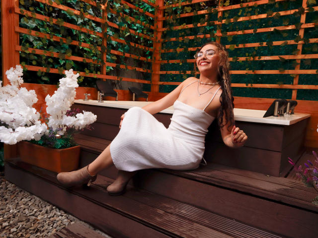 AdaraAngel with tits in a white dress, sitting on dark wooden steps, wearing brown boots, smiling, glasses on, wavy brown hair, green wall, white plant on left.