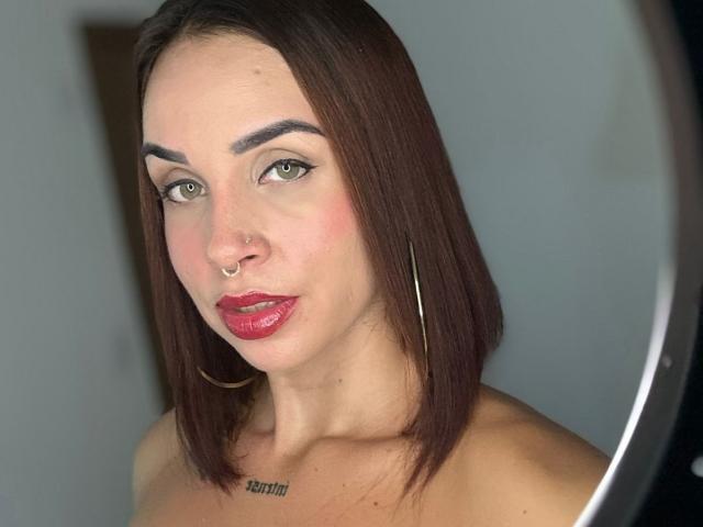 LunaX69, avec des cheveux bruns mi-longs, des yeux verts, du rouge à lèvres, un anneau au nez et des boucles d'oreilles créoles, regarde l'appareil photo. Elle a un petit tatouage sur la poitrine..