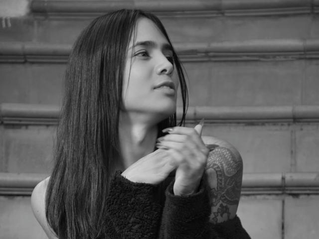 Anflamel, con cabello largo y oscuro, un brazo tatuado y un suéter negro, mirando hacia un lado, con las manos cerca del cuello, en blanco y negro. Los senos no son visibles, la vulva no es visible, el trasero no es visible..