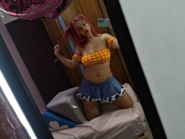 MarianneBowe with pink hair, wearing a yellow off-shoulder crop top and blue cow print skirt, kneeling on a pink bed, holding her hair, showing her tits.