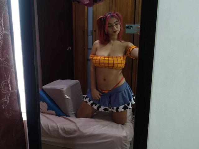 MarianneBowe with pink pigtails, glasses, orange off-shoulder top, blue cow-pattern skirt, exposing her small orange panties, kneeling on a bed, mirror selfie.