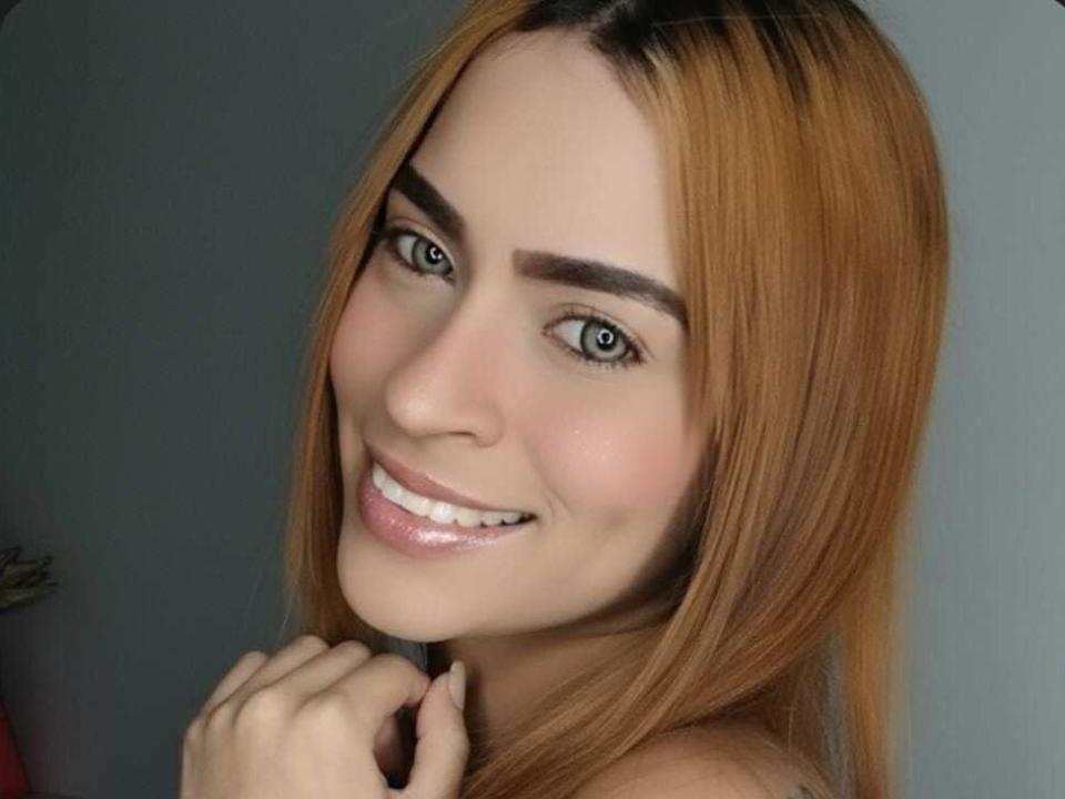ZoeParis tem cabelos castanho-claros, lisos, olhos verdes e um sorriso que mostra seus dentes brancos. Ela está tocando o queixo com a mão direita, parecendo adorável e feliz..