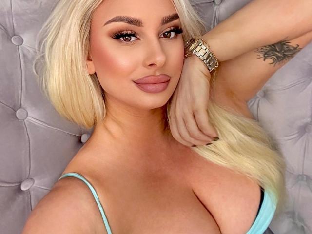 LuxuriaBlond with blonde hair, full lips, and large tits, wearing a blue top, gold watch, and a tattoo on her arm, lying on a tufted gray surface.