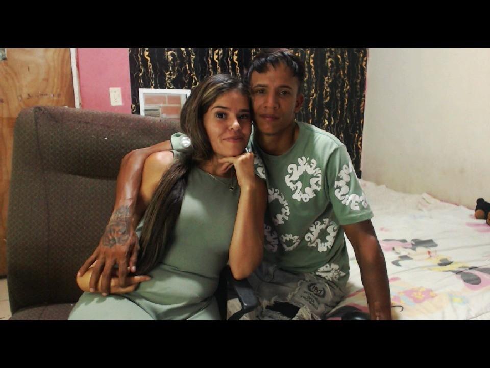 TheStarsOfLove, eine lateinamerikanische Frau mit langen, braunen Haaren in einem grünen Kleid, und ein lateinamerikanischer Mann mit Tattoos in einem grünen Hemd, sitzen auf einem Sofa, sein Arm um sie gelegt..