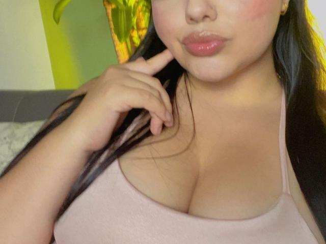 LaraJames with pouty pink lips, touching her chin, wearing a pink tank top that shows her big, round tits. She has long, dark hair and a fair skin tone.