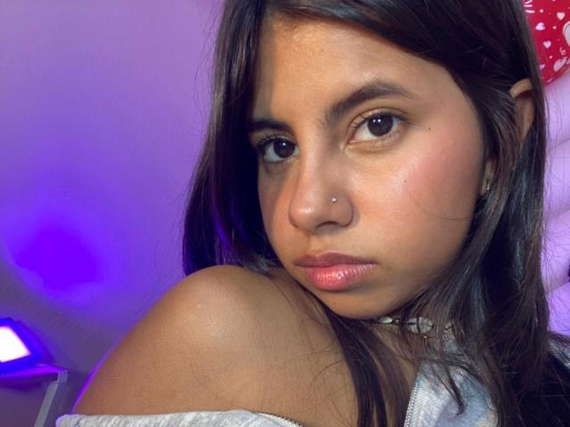 CelesteMorel has dark brown eyes, a small nose piercing, and a choker with white beads. Her brown hair is down, and she's wearing a white off-shoulder top.