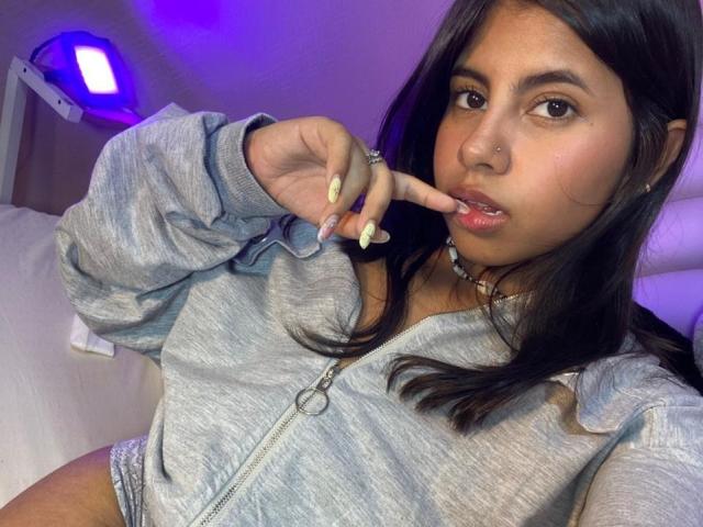 CelesteMorel with long black hair, light brown skin, and a nose ring, wearing a gray zip-up hoodie, biting her yellow-nail-manicured finger, with a cute, pouty expression.