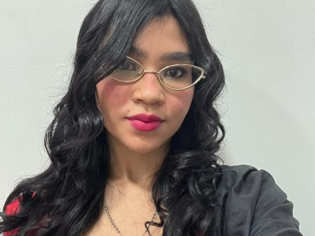 Mia Valenti, con su largo cabello negro y ondulado, gafas de montura dorada, lápiz labial rosa, una camisa negra y un collar, tiene un rostro adorable y un cutis ligeramente sonrosado..