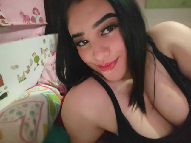 AlannaLonx, avec ses cheveux noirs, ses sourcils épais et son rouge à lèvres rouge, souriant à la caméra, portant un débardeur noir, montrant ses grandes seins, allongée sur un lit coloré..