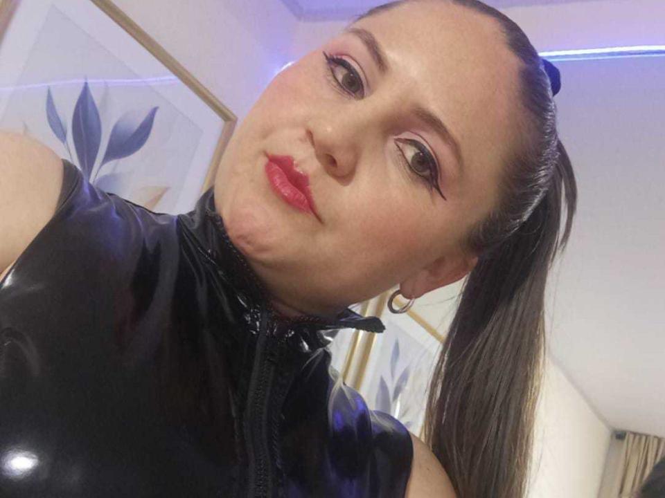 TiffanysTors mit rotem Lippenstift, schwarzem Latextop und langem, braunem Pferdeschwanz, wirkt leicht genervt, trägt Ohrringe und befindet sich in einem gut beleuchteten Raum..