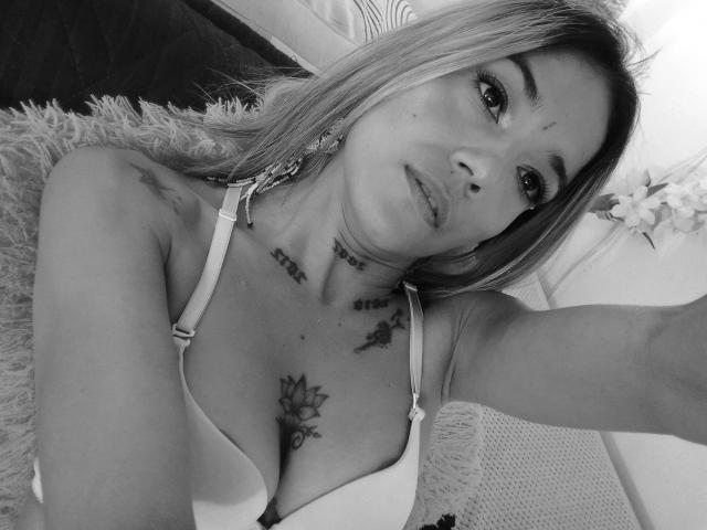 Selfie em preto e branco de Veroo69, com seios médios, tatuagens e um sutiã branco, deitada em um travesseiro felpudo, olhando para a câmera com um leve semblante de descontentamento..
