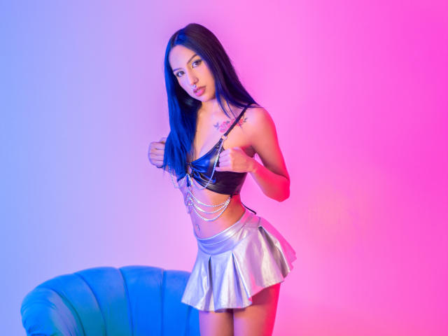 EvilWomen with long blue hair, small tits, wearing a black leather bra, silver skirt, and chain accessories, standing in pink-blue lighting, holding her bra straps.